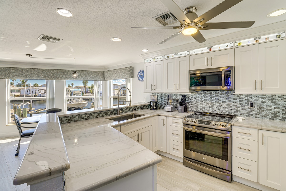 A modern kitchen features sleek white cabinetry and a spacious island with a marble countertop. Stainless steel appliances, including an oven and dishwasher, are visible. Natural light floods the area through large windows, offering views of the waterfront. A ceiling fan provides added comfort.