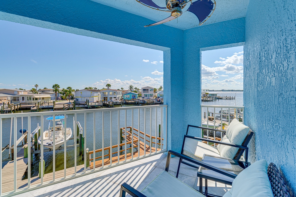 A shaded balcony is framed by two openings, revealing scenic waterfront views. Two lounge chairs are positioned to face the water, complemented by a ceiling fan overhead. The vibrant blue walls create a cheerful atmosphere while glimpses of boats and docks can be seen beyond the railing.