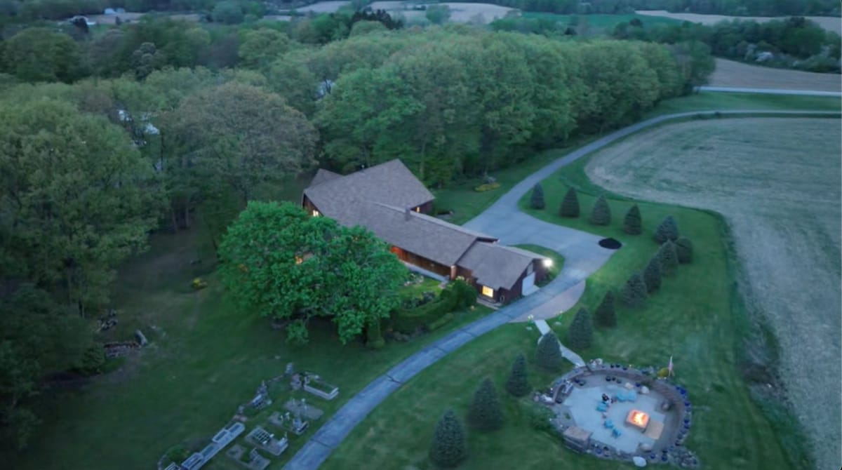An aerial view captures the expansive property of Pokey Moonshine, surrounded by lush greenery. The house features a spacious driveway leading up to it. A cozy fire pit area is visible, set in a landscaped yard, perfect for gatherings under the sky.