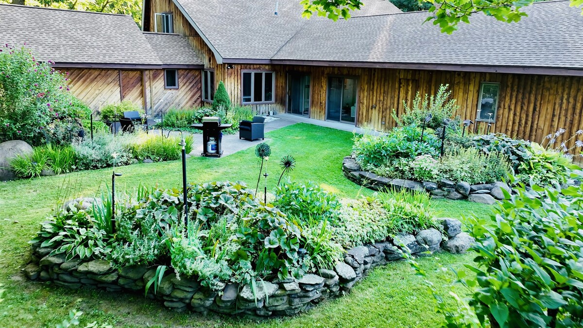 A well-maintained garden area features lush greenery and flowering plants, bordered by stone walls. The spacious lawn leads to the wooden exterior of the house, which has large glass doors and a welcoming patio area for outdoor seating.