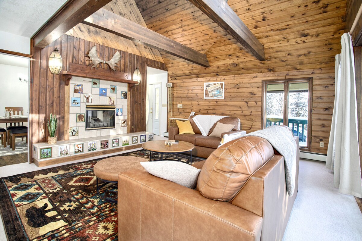 A spacious living room features elevated wooden ceilings and a warm color palette. A large leather sectional sits on a vibrant rug, while a wall-mounted fireplace is framed by a collection of photos. Natural light filters through the windows, complementing the rustic atmosphere.