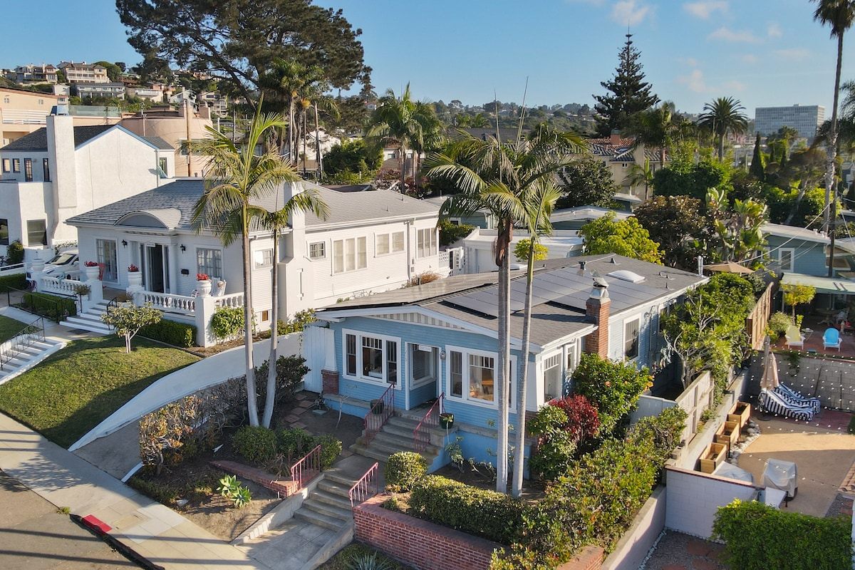 The exterior view showcases a charming single-story home with blue accents surrounded by lush greenery and palm trees. The landscaping features a well-maintained yard, accessible steps leading to the entrance, and neighboring homes adding to the inviting neighborhood ambiance.