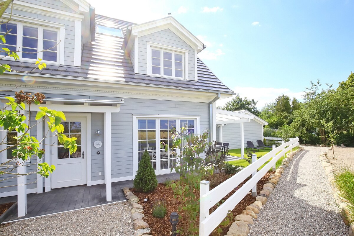 A light-colored, two-story house with a sloped roof is set amidst greenery. A welcoming front porch is visible, along with a pathway lined by stones. Comfortable outdoor seating is seen in the garden area, promoting an inviting appearance.