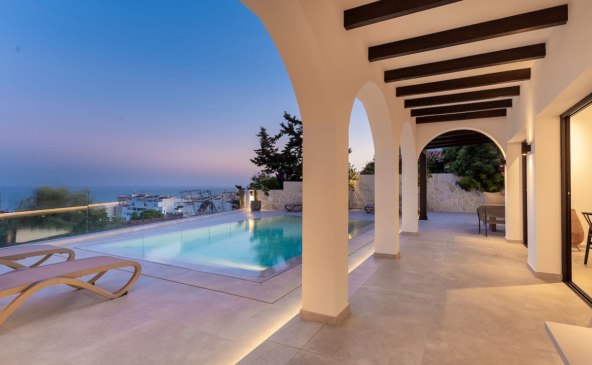 An outdoor terrace is framed by elegant arches, offering a view of the tranquil pool. Sun loungers are positioned beside the shimmering water, with soft evening light casting a serene ambiance. Lush greenery is visible in the background, enhancing the peaceful setting.