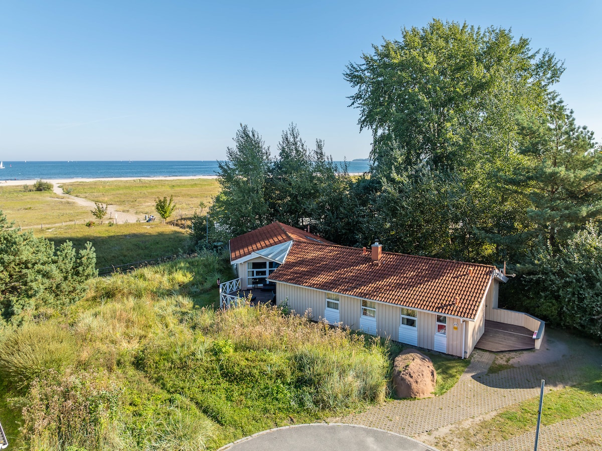 A single-story vacation house is set amidst lush greenery, with a red-tiled roof and light-colored exterior. Large windows allow for natural light, and a path leads to the entrance. The nearby beach and expansive sea view are visible in the background.