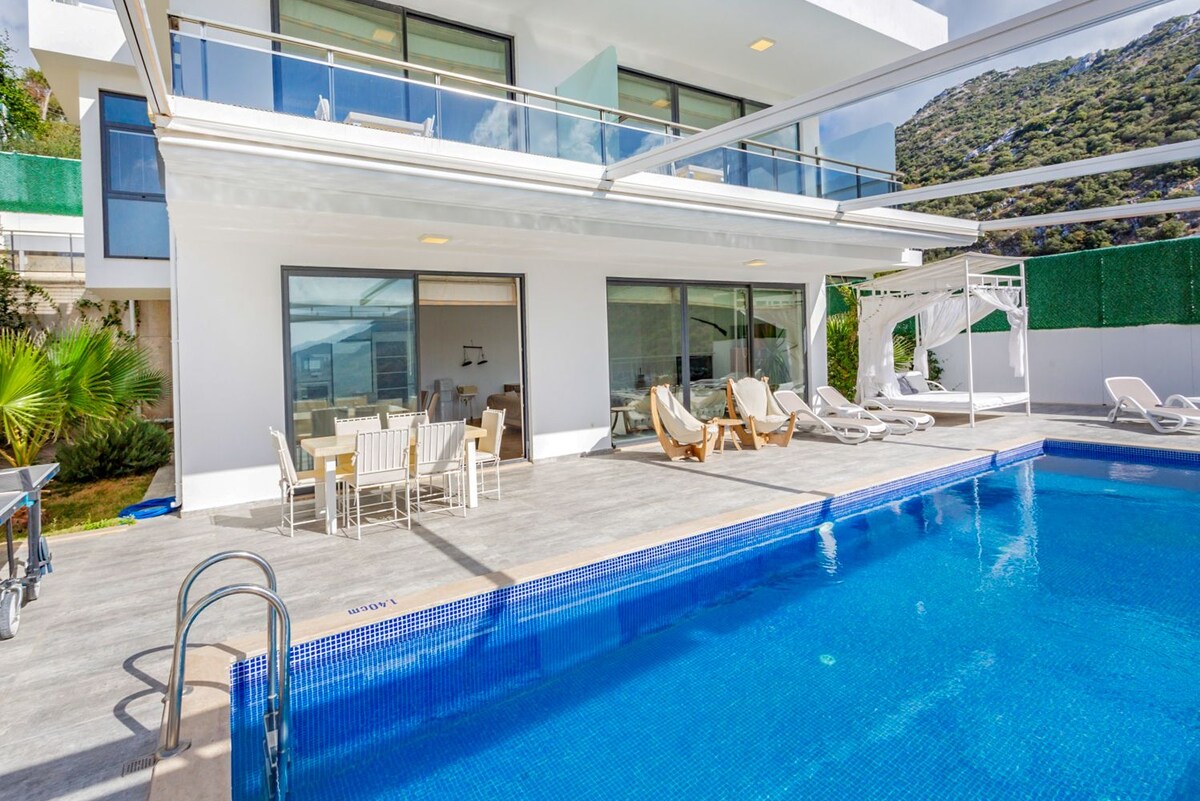 A spacious outdoor area features a large swimming pool surrounded by sun loungers and a double day bed. A dining table with chairs is positioned on the terrace, offering views of the villa's modern architecture and the lush landscape beyond.
