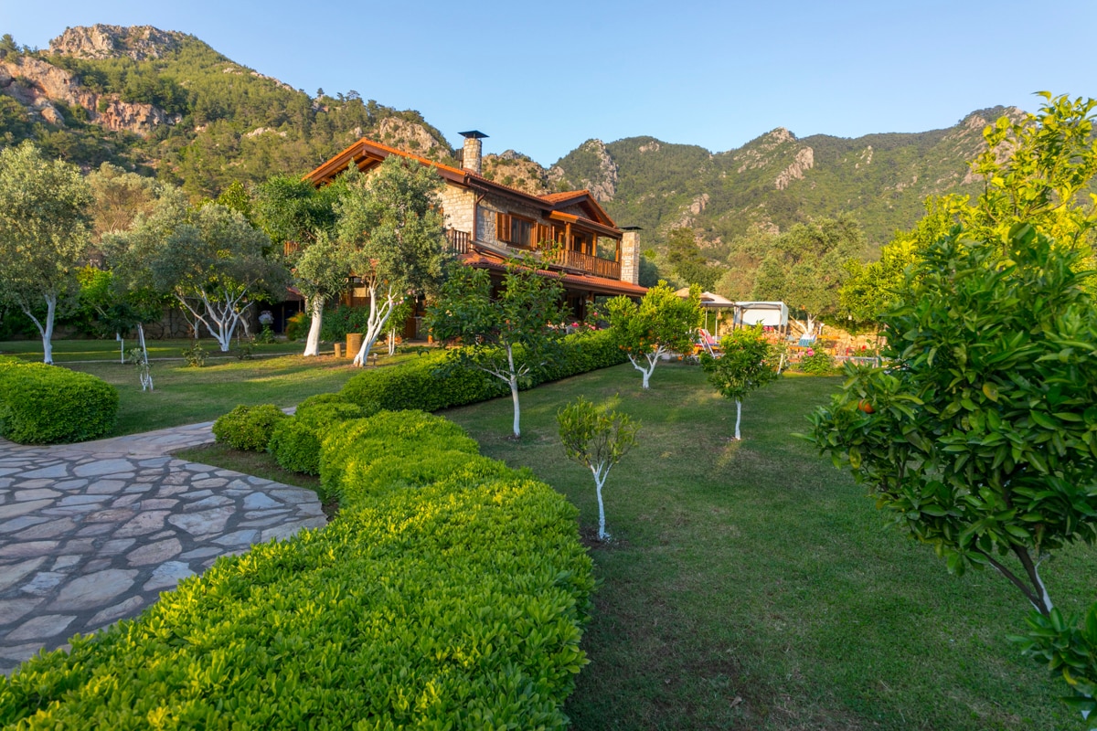 A spacious villa is nestled among lush gardens, featuring neatly trimmed hedges and fruit trees. The stone exterior and wooden accents of the house harmonize with the natural landscape. Majestic mountains are visible in the background, providing a serene backdrop.