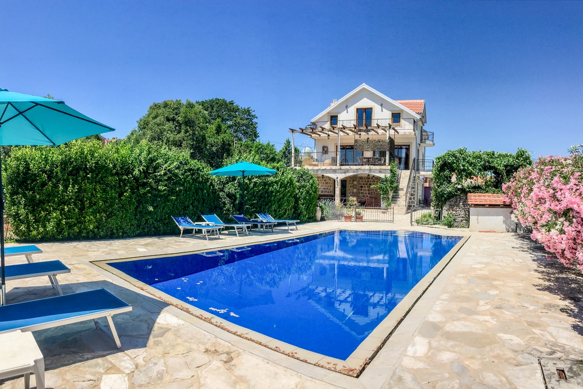 A private outdoor space features a spacious blue pool surrounded by sun loungers. Bright umbrellas provide shade, while lush greenery and flowering shrubs enhance the setting. The villa stands in the background, showcasing a multi-level design with a covered terrace and abundant natural light.