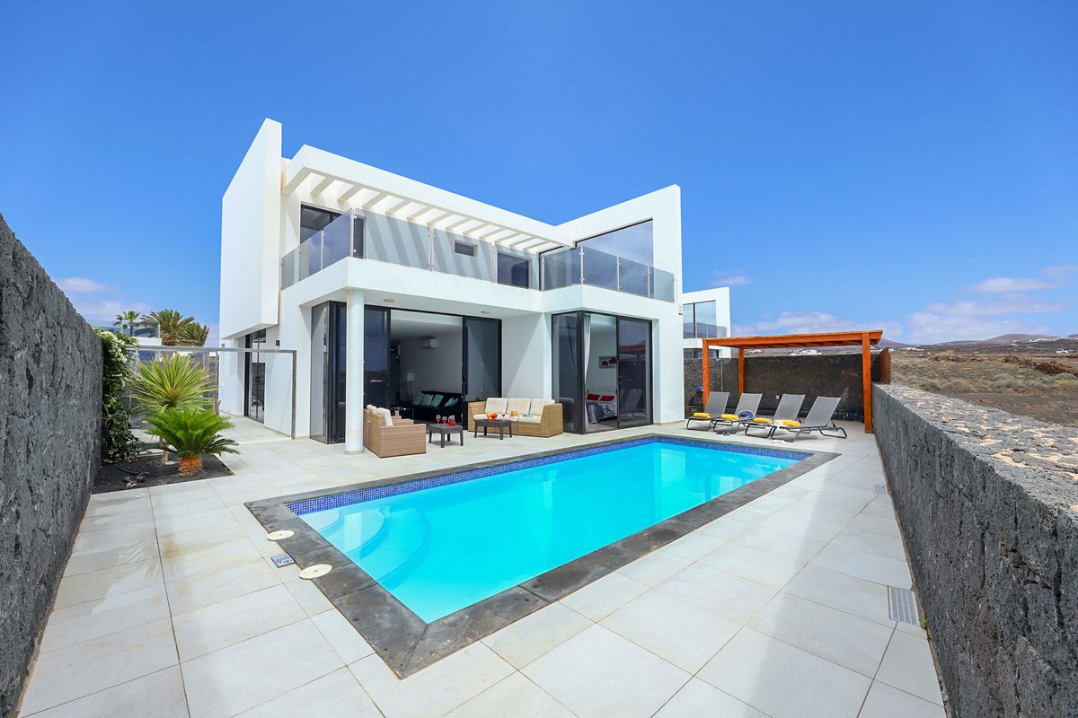 A modern two-story villa is showcased beside a private heated swimming pool. The white exterior contrasts with the clear blue sky, while outdoor seating areas are arranged to encourage relaxation. A patio area is visible, providing a welcoming space for outdoor gatherings.