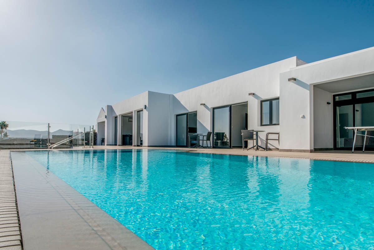 The villa features a spacious infinity pool with crystal-clear water, surrounded by a sunlit terrace. Large glass doors open to the indoor living spaces, enhancing the connection with the surrounding landscape. A vibrant blue sky complements the modern white architecture.