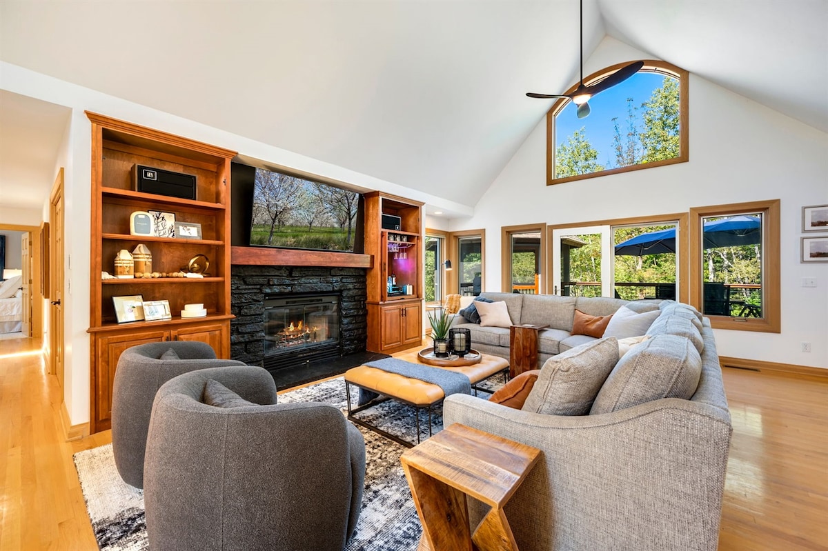 The living room features a spacious sectional sofa arranged around a coffee table, with a cozy farmhouse-style fireplace as the focal point. Large windows allow natural light to fill the space, offering views of the surrounding woodland. Custom cabinetry adds warmth and charm.