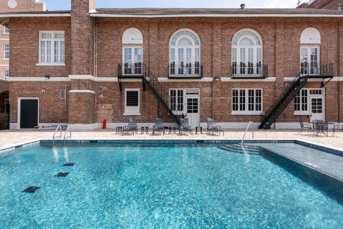A refreshing swimming pool is situated in front of a brick building featuring large arched windows. Comfortable loungers are placed around the pool, with stairways leading up to balconies. The clear water reflects the sunlight, creating a tranquil atmosphere.