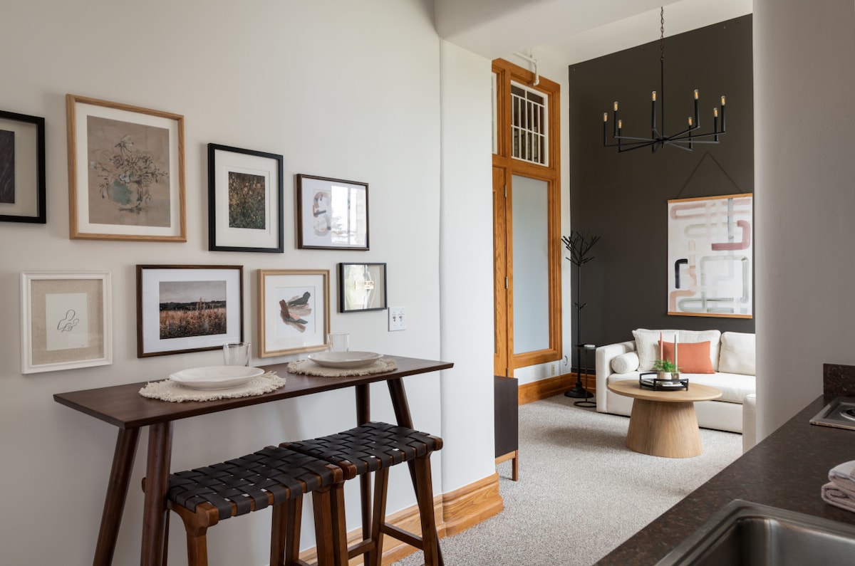 A dining area is presented with a wooden table and two woven stools, complemented by framed artwork on the wall. The open layout leads to a living space featuring a couch and additional decorative elements, reflecting a contemporary yet cozy aesthetic.