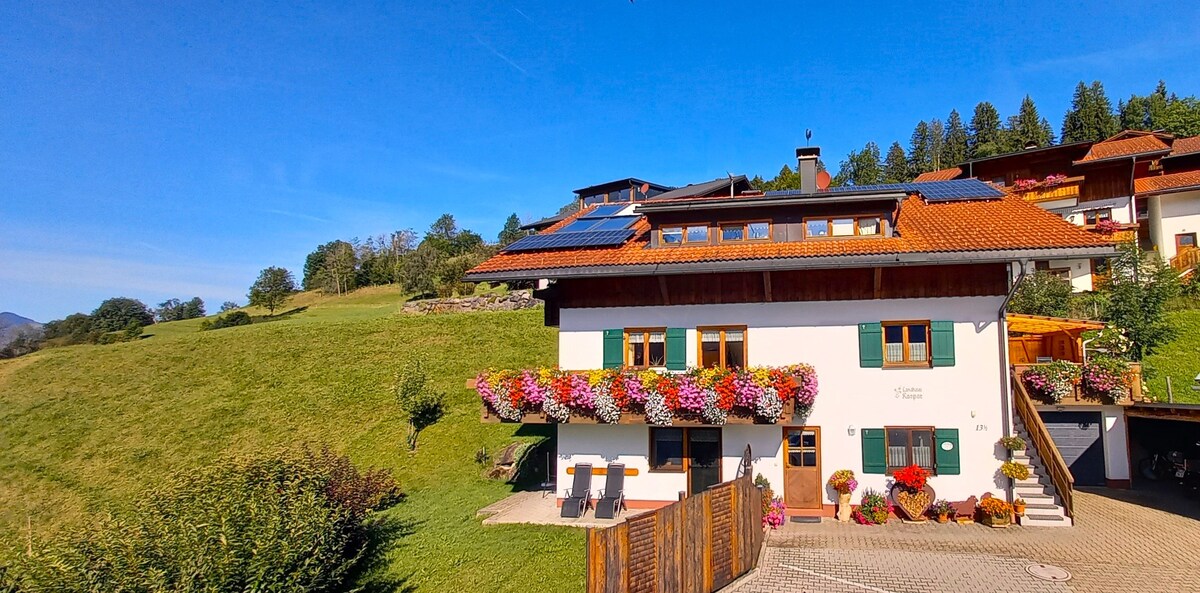The exterior of a two-story holiday apartment building showcases a vibrant display of blooming flowers along the windows. A well-maintained lawn and hillside are visible in the background, under a clear blue sky. Solar panels are installed on the roof, indicating a sustainable energy use.