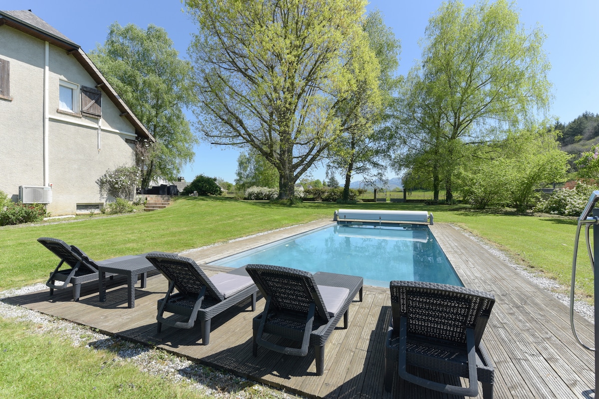 An outdoor area presents a heated swimming pool surrounded by wooden decking. Several lounge chairs are arranged nearby, set within a green lawn adorned with trees. The tranquil atmosphere is enhanced by the clear sky and natural surroundings.
