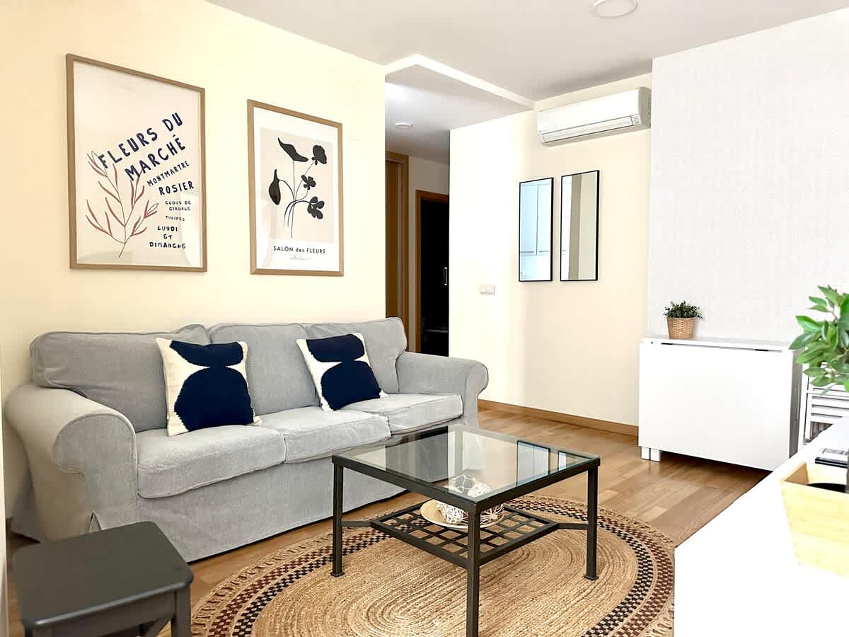 A comfortable living area is highlighted by a gray sofa adorned with decorative pillows. A glass coffee table sits atop a round woven rug, creating a cozy seating arrangement. Artwork is displayed on the wall, while a small potted plant adds a touch of greenery to the space.