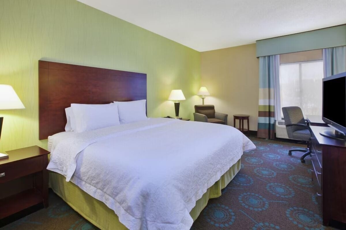 A spacious hotel room features a king bed with crisp linens and patterned carpeting. Two bedside lamps provide soft lighting. A seating area includes a chair ottoman, work desk, and television, with a window dressed in sheer and patterned curtains allowing natural light.