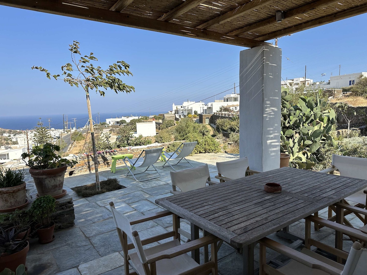 A spacious outdoor veranda features a wooden dining table surrounded by eight chairs, offering panoramic views of the Aegean Sea and nearby villages. Sun loungers are positioned nearby, and potted plants add a touch of greenery to the stone patio.