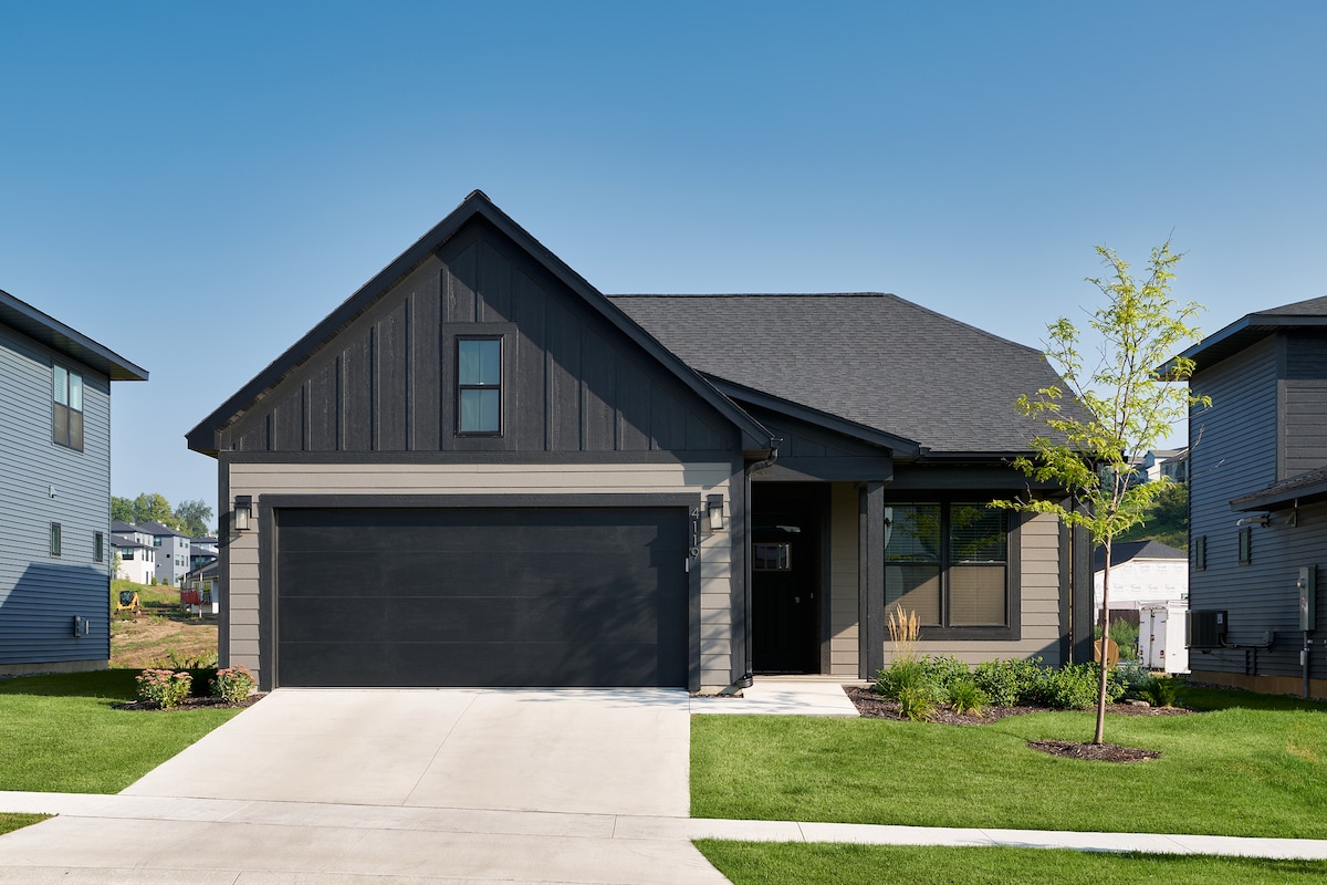 A modern single-family home showcases a dark exterior with a spacious two-car garage. The front yard is neatly landscaped, featuring green grass and a young tree. A covered porch provides shaded entry, with large windows allowing natural light to stream indoors.