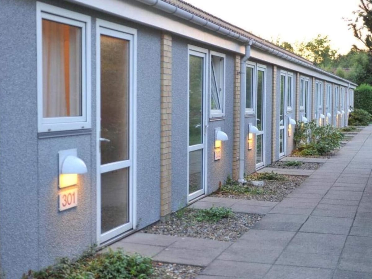 The exterior of the holiday home displays a series of neatly arranged rooms, each with a distinct front door. Soft outdoor lighting illuminates the pathway, lined with well-maintained shrubs and small stones. Windows adorned with simple curtains allow glimpses of the interior.