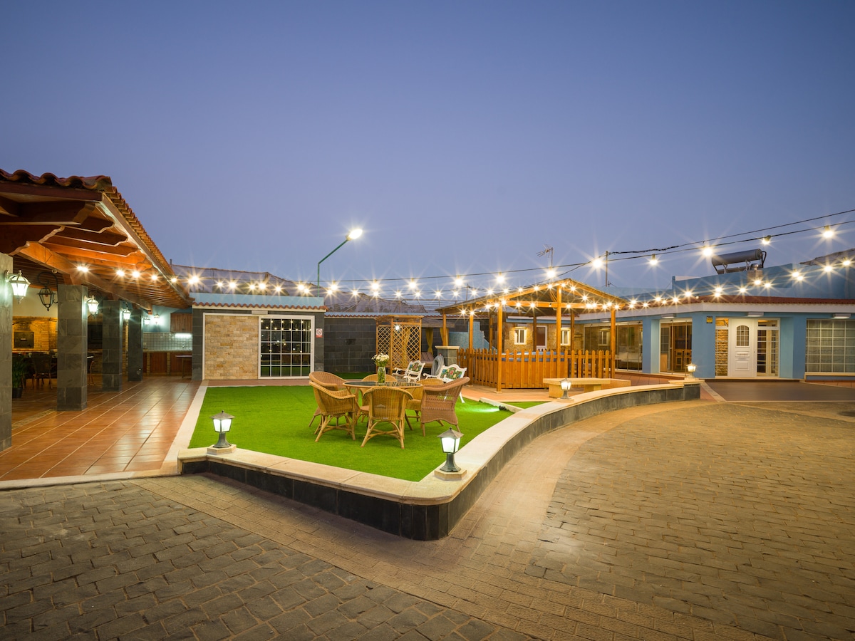 An outdoor area is illuminated by string lights, creating a welcoming atmosphere. A circular patio features comfortable seating arrangements made of wood, surrounded by manicured grass. The adjacent structure showcases a blend of stone and traditional architecture, with multiple windows reflecting the soft evening light.