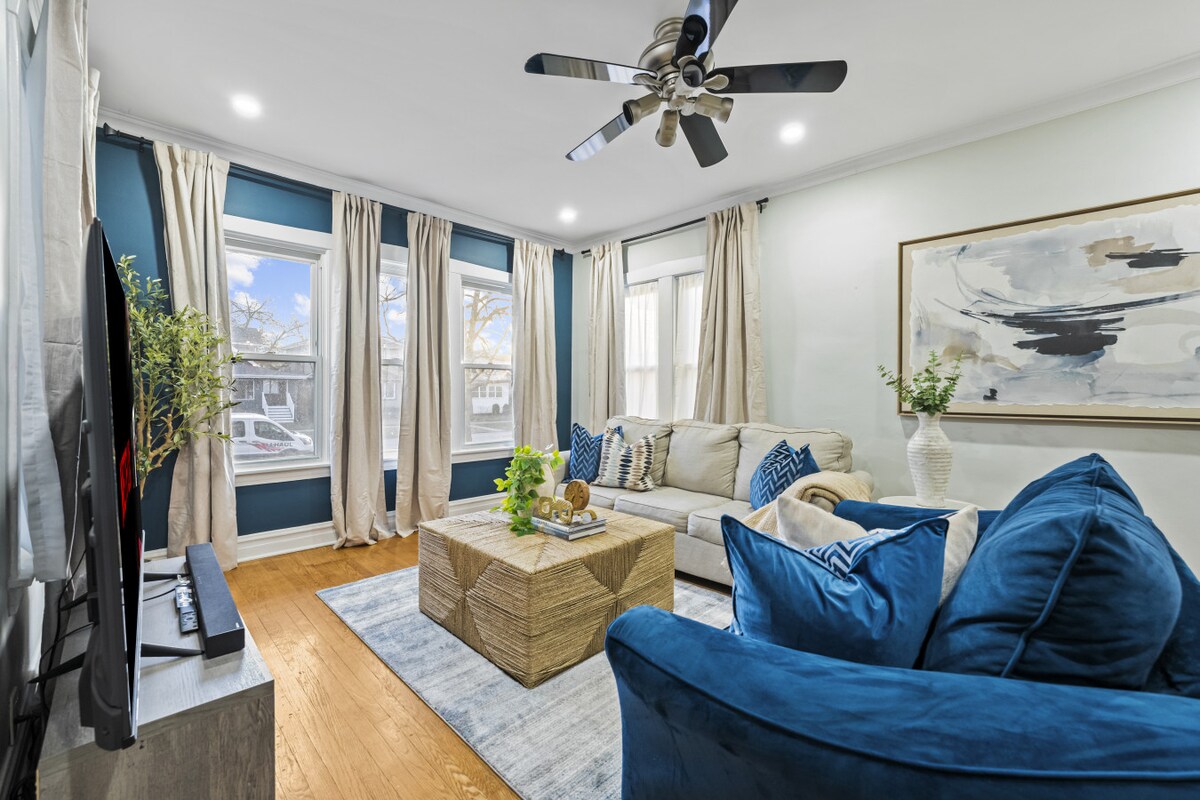 A comfortable living area is featured with a sectional sofa adorned in cream and blue hues. Natural light floods in through two large windows, framed by soft curtains. A light wooden coffee table anchors the space, accompanied by a vibrant blue armchair and decorative plant arrangements.