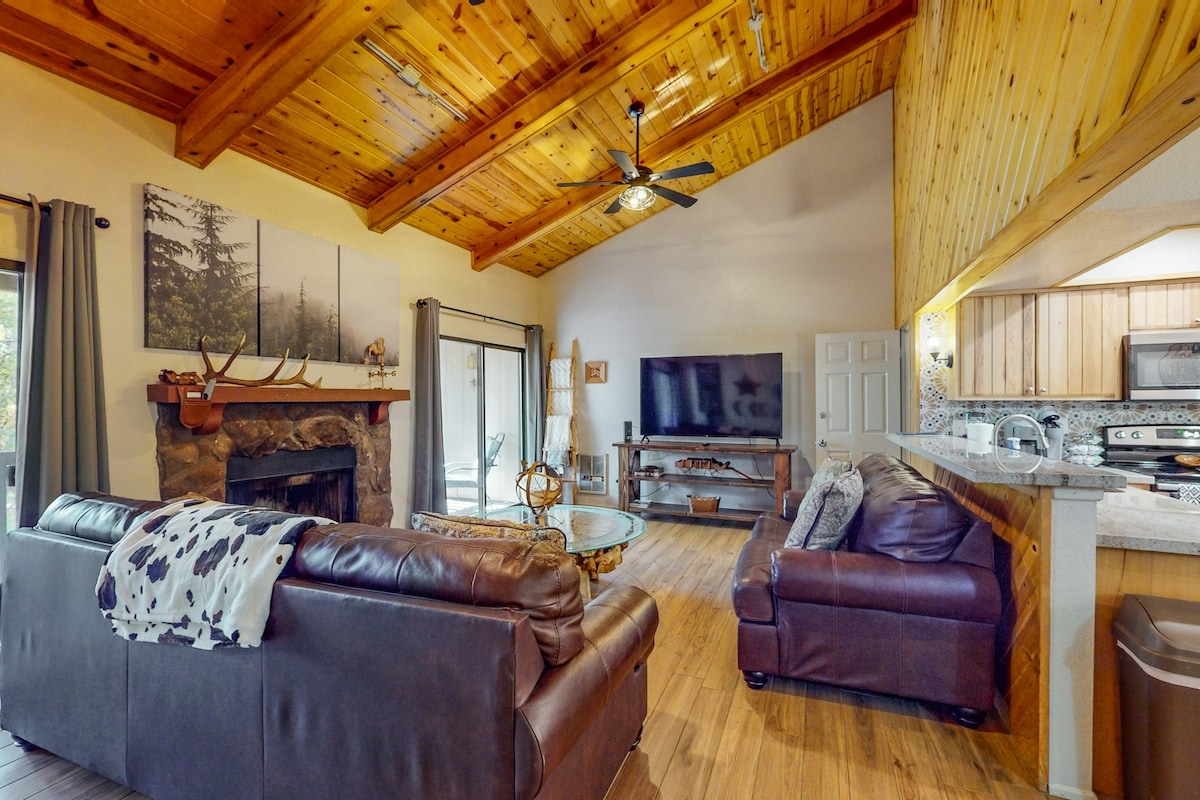 The living area features wood-paneled vaulted ceilings and a cozy wood-burning fireplace. Comfortable leather seating surrounds a glass coffee table, while a television is strategically placed for easy viewing. Large windows allow natural light to fill the space, enhancing the inviting layout.