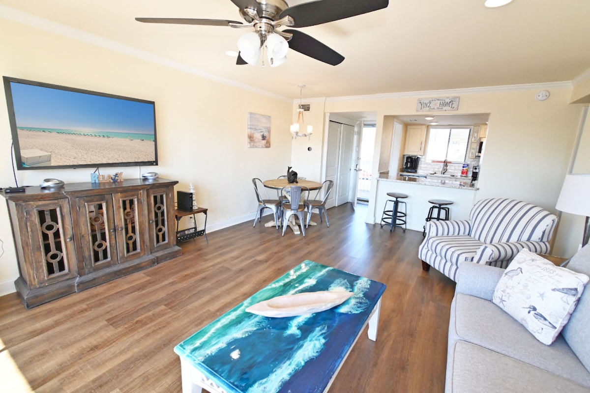 The living room features a comfortable couch and a large wall-mounted Smart TV, displaying scenic beach imagery. A colorful coffee table and a detailed wooden entertainment unit add character. A dining area for four is visible, along with the entrance to the kitchen.