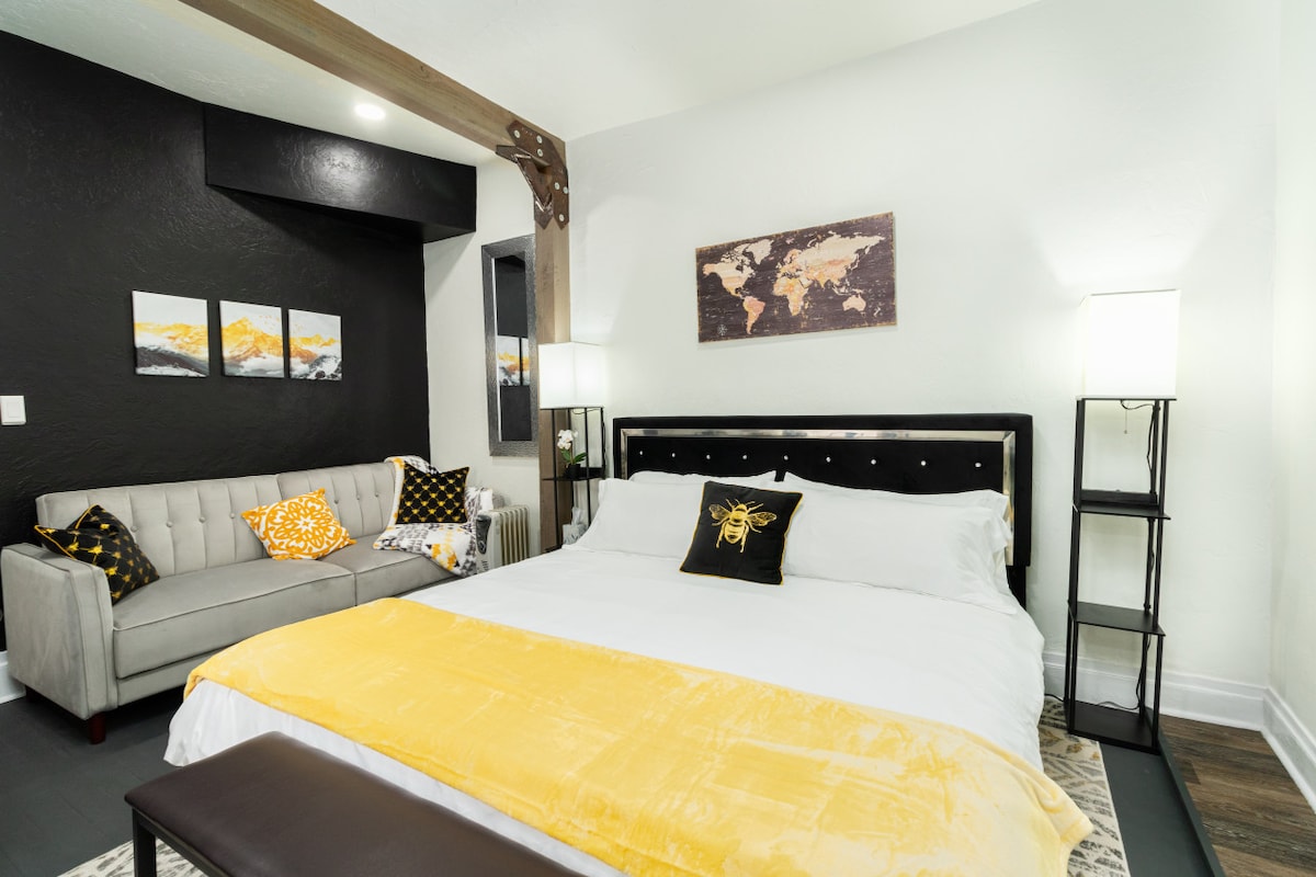 A spacious sleeping area contains a plush king-sized bed dressed in crisp white linens, accented by a vibrant yellow throw. A comfortable gray sofa with decorative pillows complements the space, while soft lighting is provided by stylish bedside lamps.