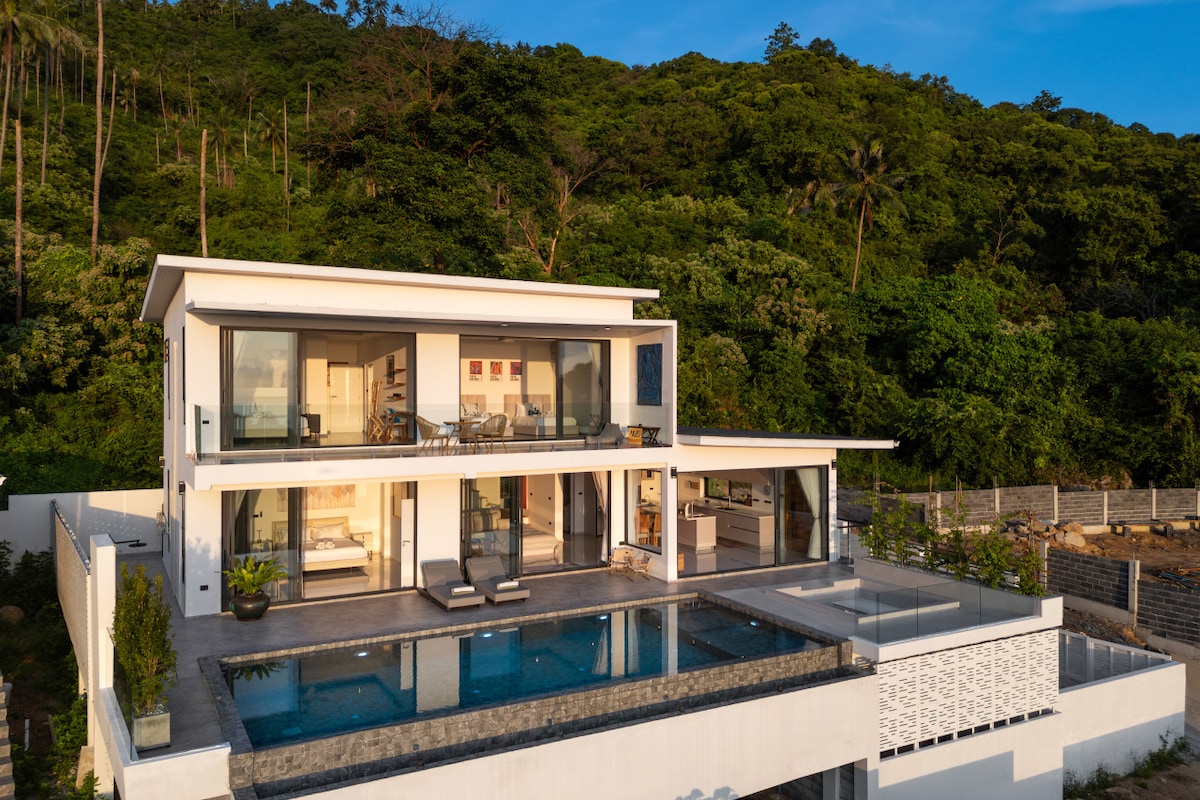 The villa's modern architecture is showcased with multiple levels, featuring large glass windows and sliding doors that provide unobstructed views of the surrounding greenery. A saltwater infinity pool extends from the main terrace, with sun loungers placed alongside for relaxation.