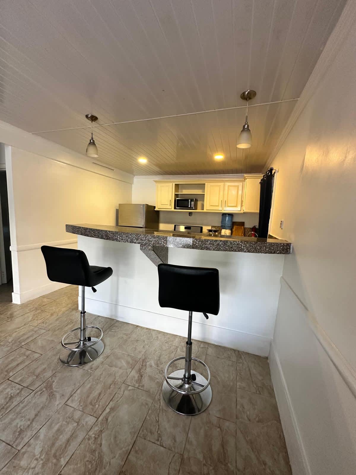 A modern kitchen area is highlighted by a sleek bar counter with two black stools. Warm overhead lights illuminate the space, while cabinetry and a microwave are visible in the background. The flooring features light-coloured tiles, contributing to a bright and spacious atmosphere.