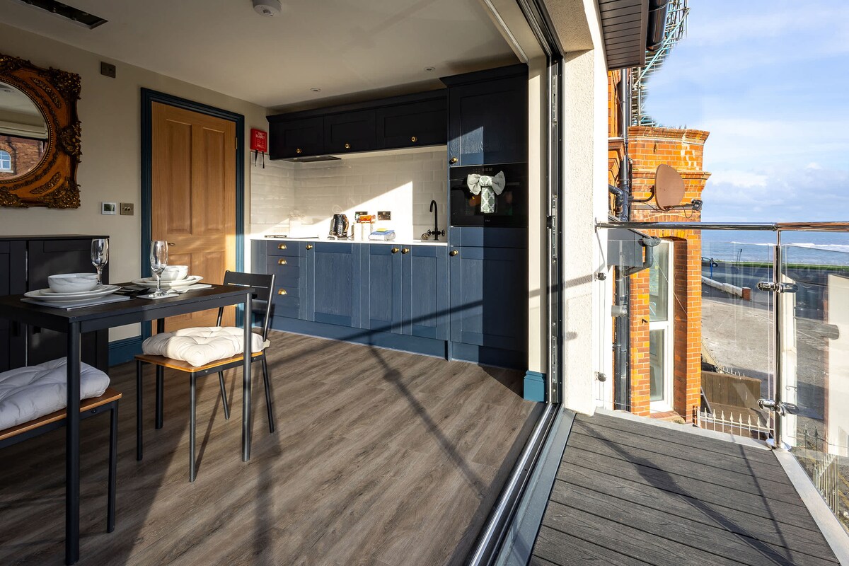 A spacious kitchen diner features modern navy cabinetry and sleek appliances, including a kettle and countertop. A dining table with black chairs is positioned near bi-fold doors, which open onto a balcony. Natural light floods the space, highlighting the stylish finishes and warm wood flooring.
