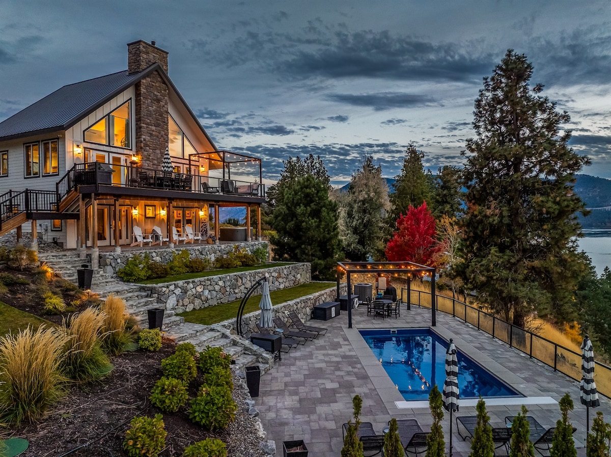 A lakeside property features a spacious outdoor area with a saltwater pool, surrounded by landscaped gardens and seating. The home's large windows reflect evening light, while panoramic views of the lake and mountains are visible in the background. A covered patio offers additional space for relaxation.