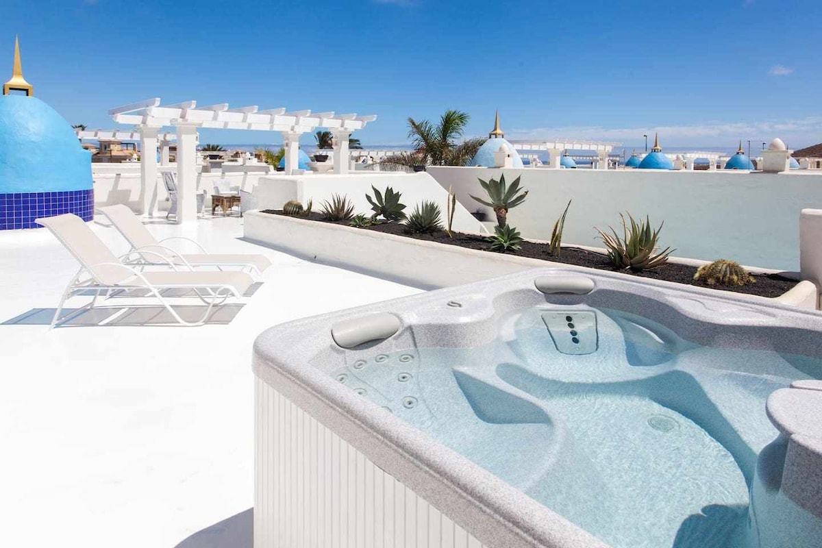 A rooftop terrace is featured, showcasing a private Jacuzzi surrounded by contemporary landscaping. Sun loungers are positioned nearby, providing a relaxing space. The bright sky and blue accents complement the tranquil environment.