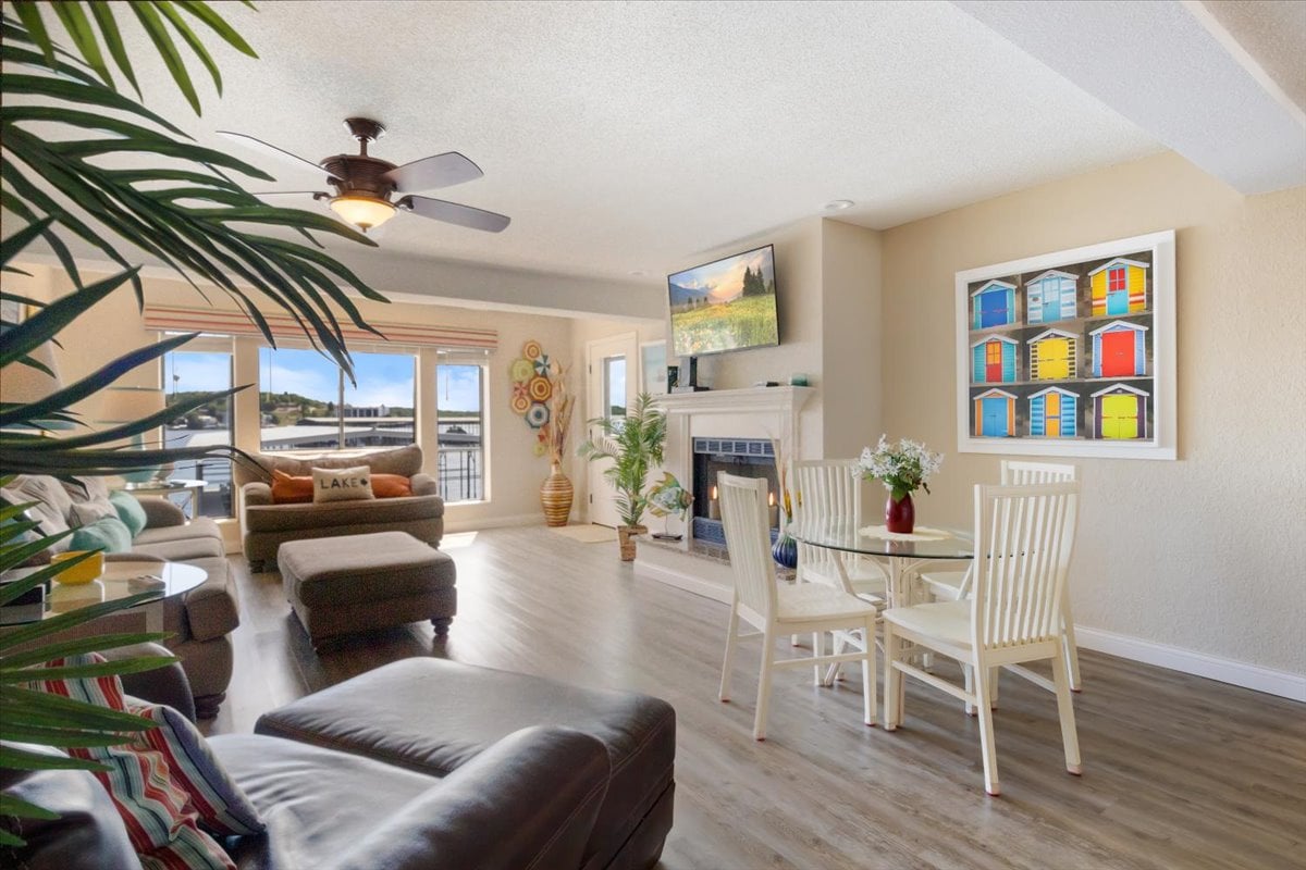 A spacious living area features a comfortable sectional sofa and cozy chairs, creating a welcoming environment. Natural light fills the room through large windows, revealing lake views. A dining table accommodates four, while colorful wall art adds a touch of vibrancy to the neutral decor.