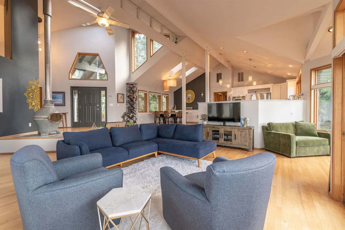 A spacious common area features a large sectional sofa and two comfortable armchairs, inviting relaxation. Large windows provide natural light and views of the outdoors. A dining table is visible in the background, accompanied by a 50-inch Smart TV on a media console.