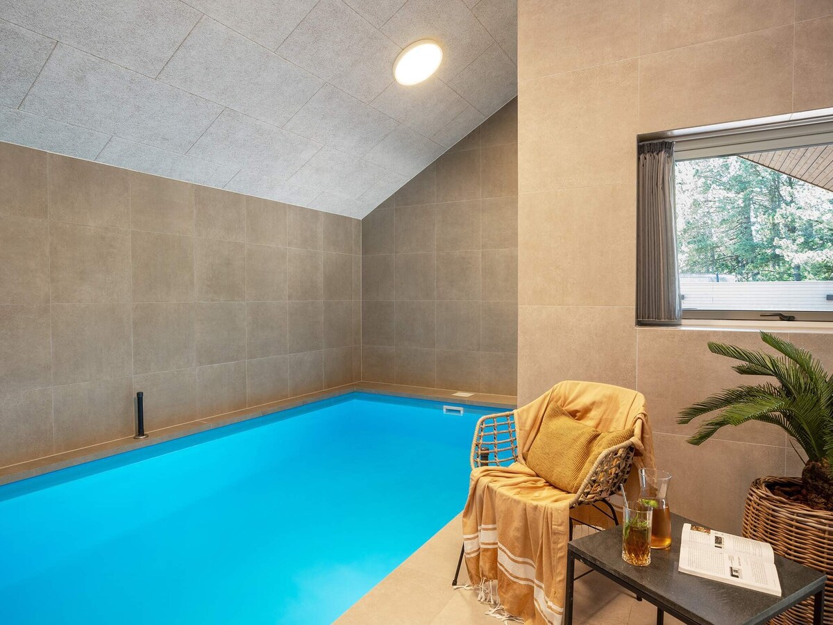 An indoor swimming pool is highlighted by smooth grey tiles and a gently sloping ceiling. A comfortable chair, draped with a light blanket, is positioned beside the pool, which features clear blue water. A refreshing drink rests on a side table, adding a touch of relaxation.