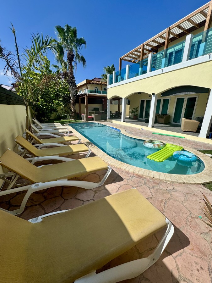 A private pool area is surrounded by tall palm trees, with several sun loungers positioned along the edge. A turquoise pool extends invitingly, complemented by a colorful float. The outdoor space is finished with a textured stone patio, enhancing the retreat's relaxing environment.
