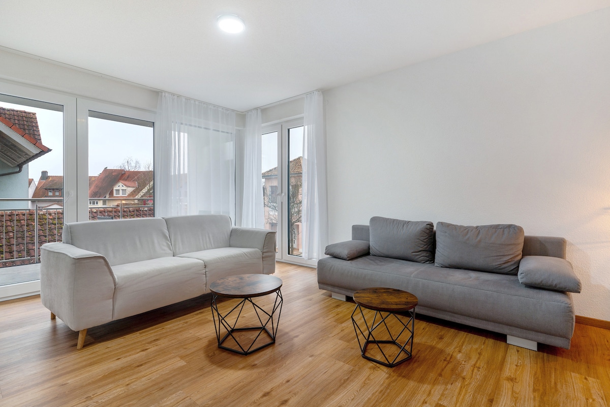 The living room features two sofas, one in light fabric and one in a darker hue. A pair of round coffee tables with geometric bases occupies the center. Large windows allow natural light to fill the space, complemented by light-colored walls and wooden flooring.