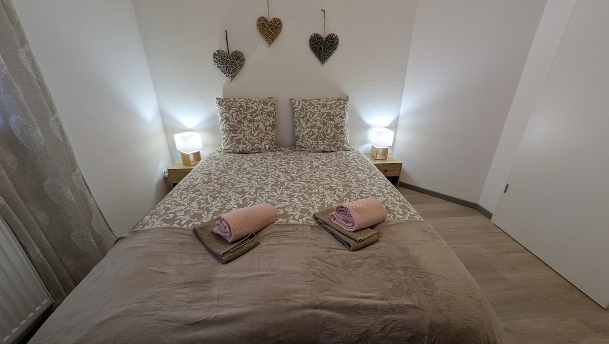 A cozy bedroom is presented with a spacious bed dressed in neutral tones, featuring patterned headboards and two bedside lamps. Soft towels are neatly arranged at the foot of the bed, creating a welcoming and organized appearance. Light gently illuminates the room from both sides.
