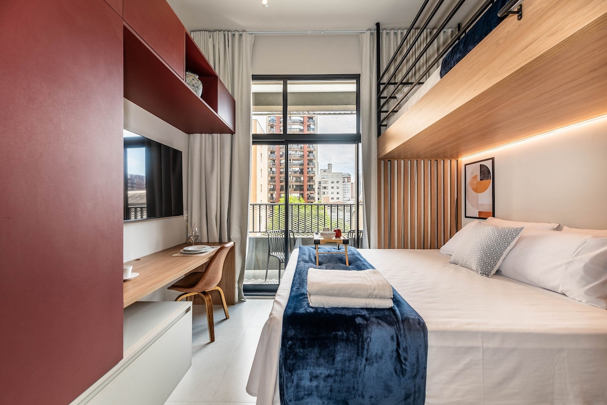A modern studio features a queen bed and a lofted bunk bed along the wooden wall. A compact workspace and a television are included. Natural light fills the space through the large window, which opens to a balcony overlooking the surroundings.