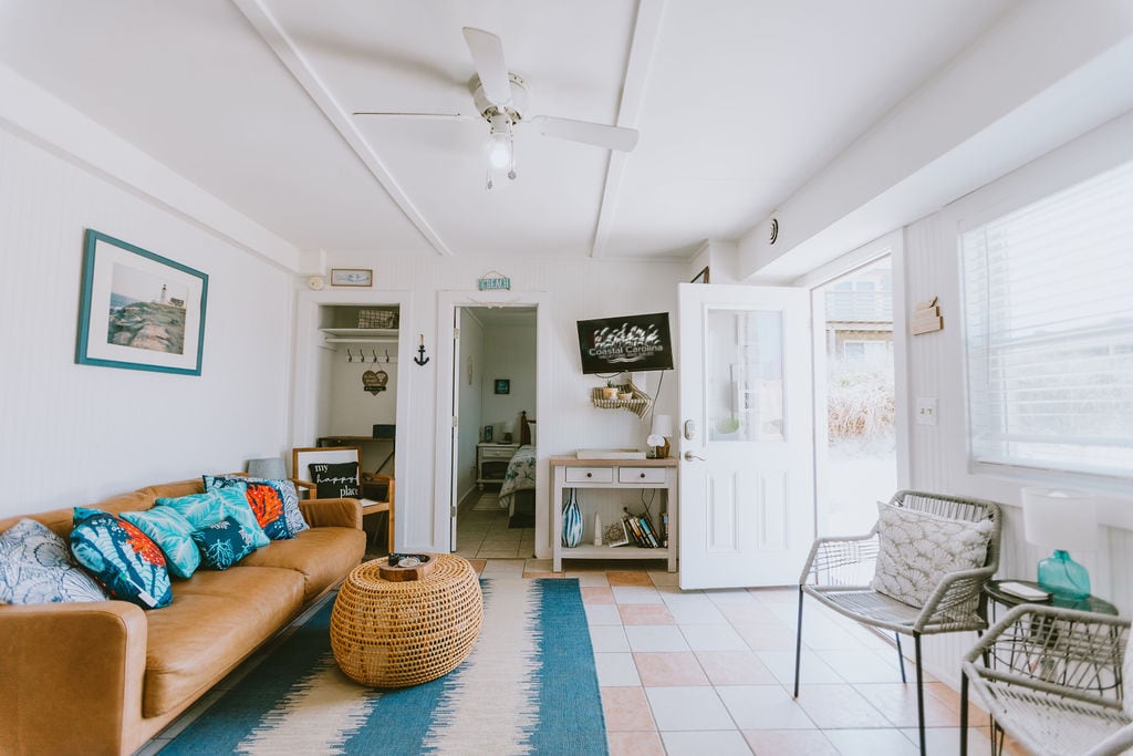 A light-filled living area features a comfortable brown sofa adorned with colorful cushions. A woven coffee table sits in front, complemented by two stylish chairs. The space is enhanced by a ceiling fan and large windows, inviting natural light and ocean breezes.