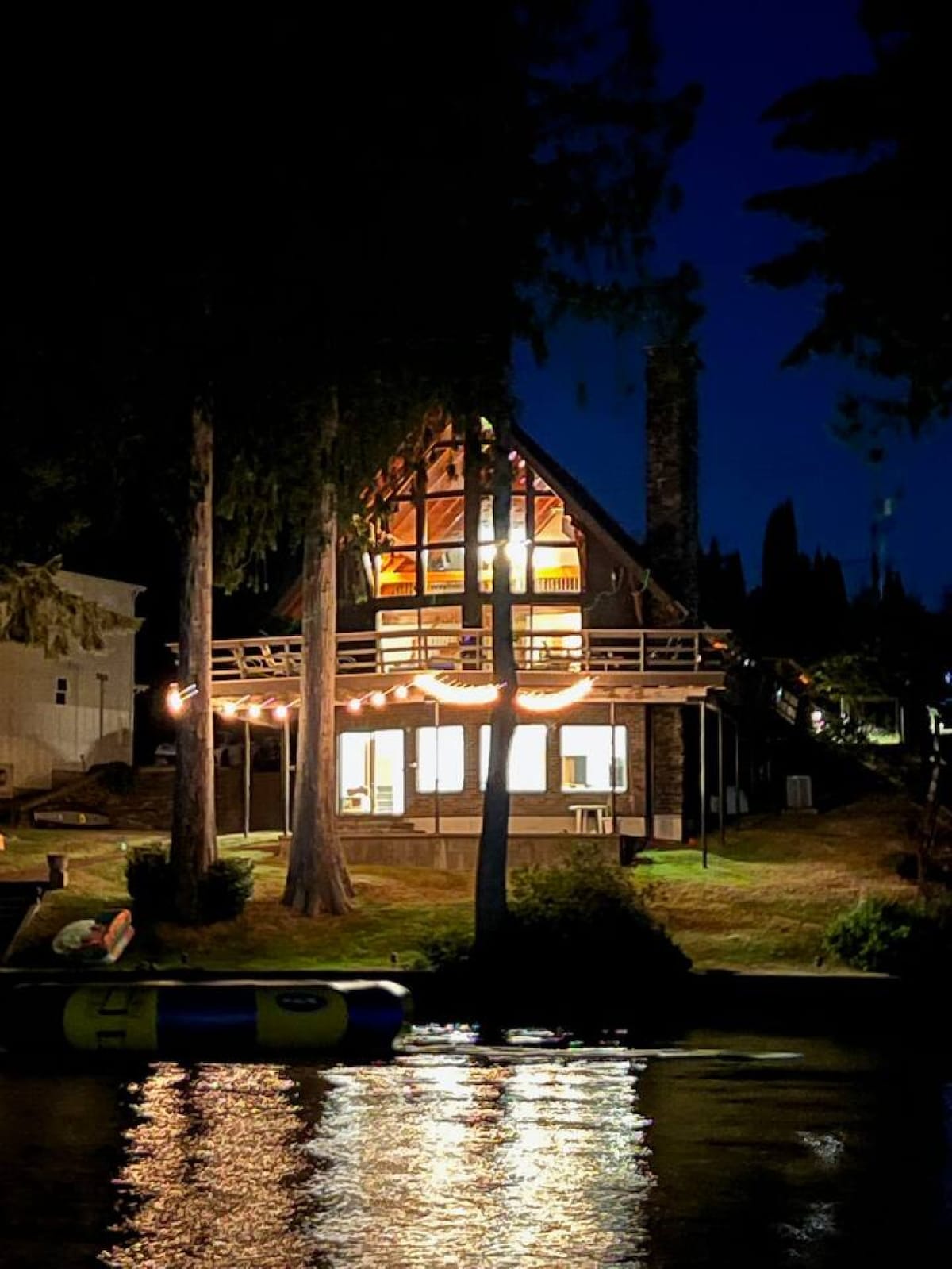 The A-frame cabin is illuminated at night, showcasing its warm glow through expansive windows. The deck, adorned with lights, overlooks the water, while tall trees frame the structure, enhancing the serene lakeside setting.