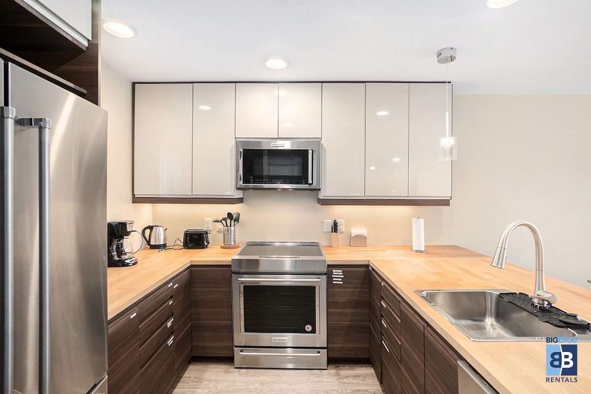 A modern kitchen features sleek cabinetry with a combination of light and dark finishes. Stainless steel appliances, including an oven and microwave, are integrated into the design. Wooden countertops provide a warm contrast, while under-cabinet lighting enhances the workspace.