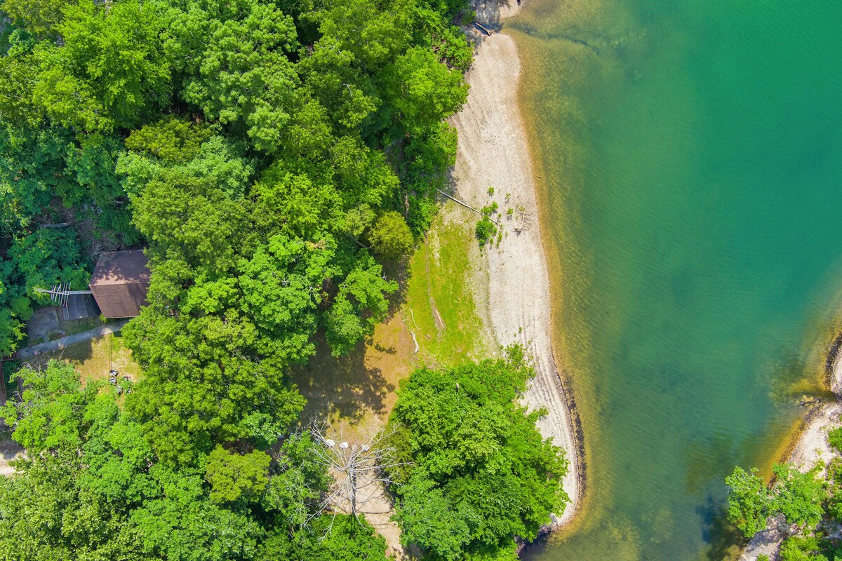 An aerial view captures a secluded beach area along Beaver Lake, surrounded by dense green foliage. The shoreline features a mix of sandy and grassy patches, leading to clear water that reflects the vibrant greenery, creating a serene natural environment.