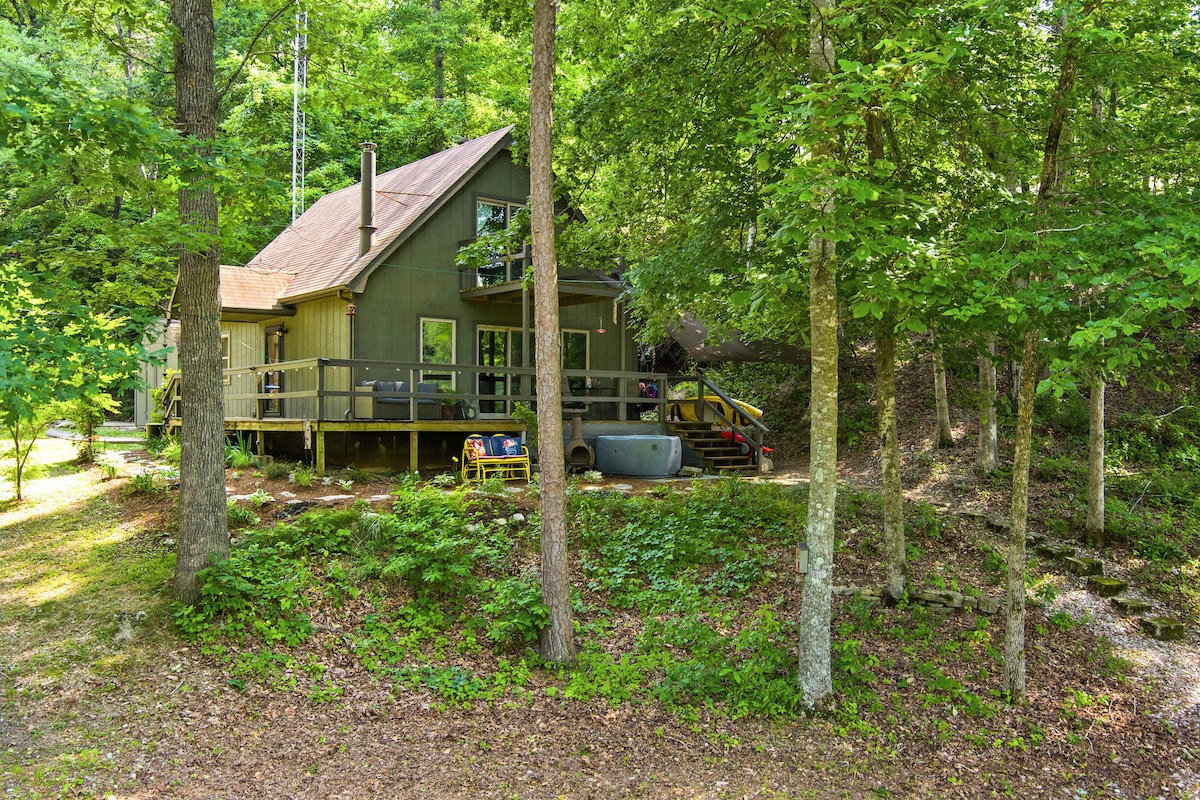 A cabin nestled among lush greenery features a large deck for outdoor relaxation. Steps lead from the deck down into the forested area, emphasizing the cabin's natural surroundings. The setting provides a serene atmosphere with ample tree coverage and gentle landscape variations.