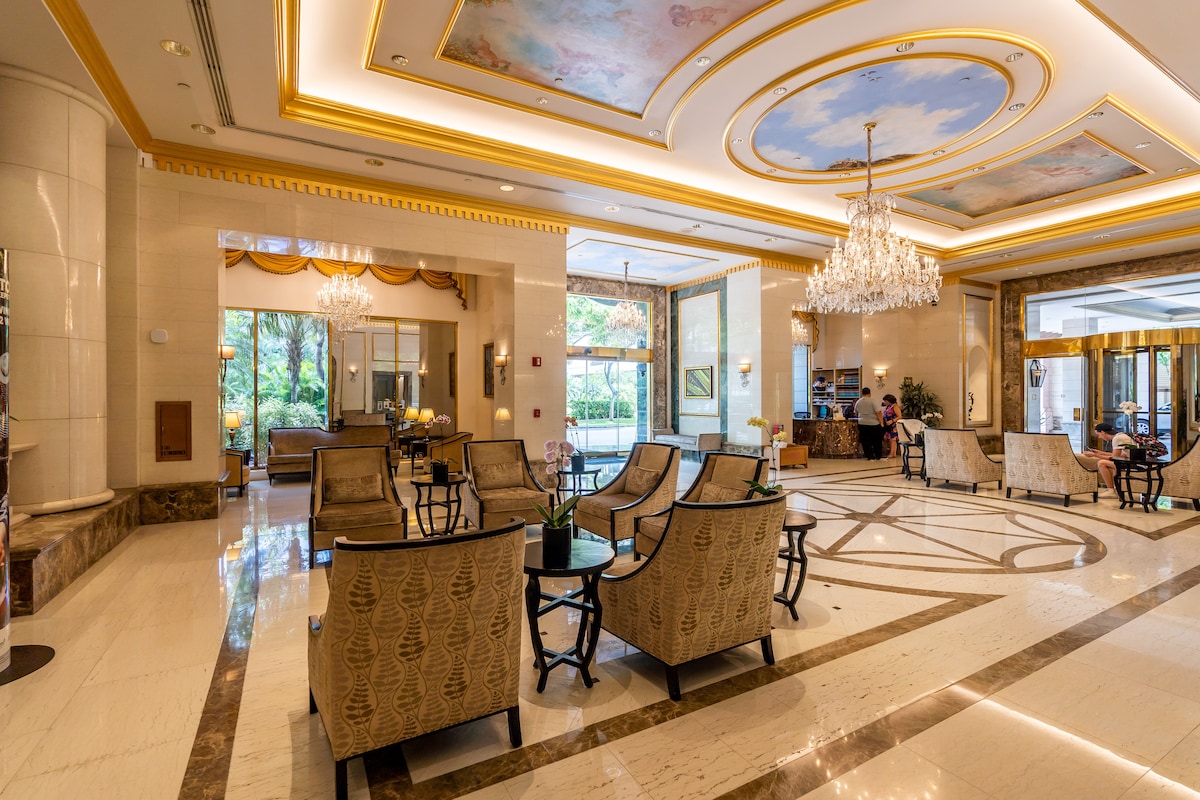 A chic lobby features elegant furnishings with tasteful armchairs arranged around small tables. Crystal chandeliers hang from the ceiling, complementing the soft lighting. Large windows allow natural light to illuminate the space, with lush greenery visible outside.