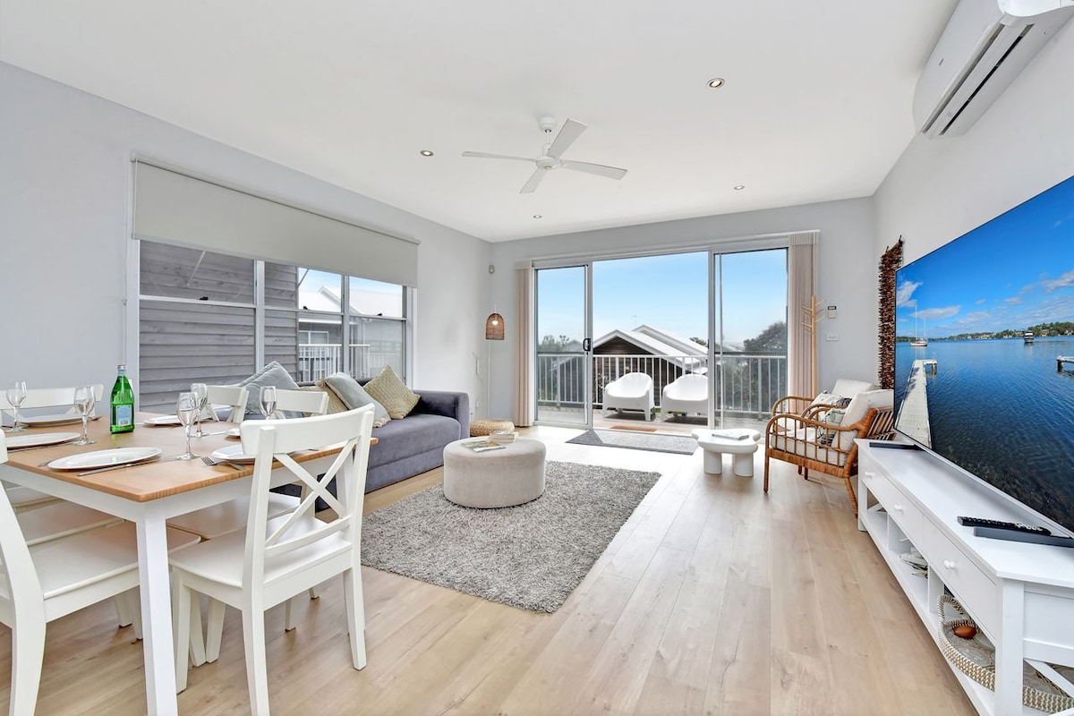 An inviting open-plan living area is featured, with a comfortable couch and a plush round ottoman. A dining table with white chairs accommodates up to four people. Large glass doors lead to a balcony, while a flat-screen TV is positioned on a white console against the wall.