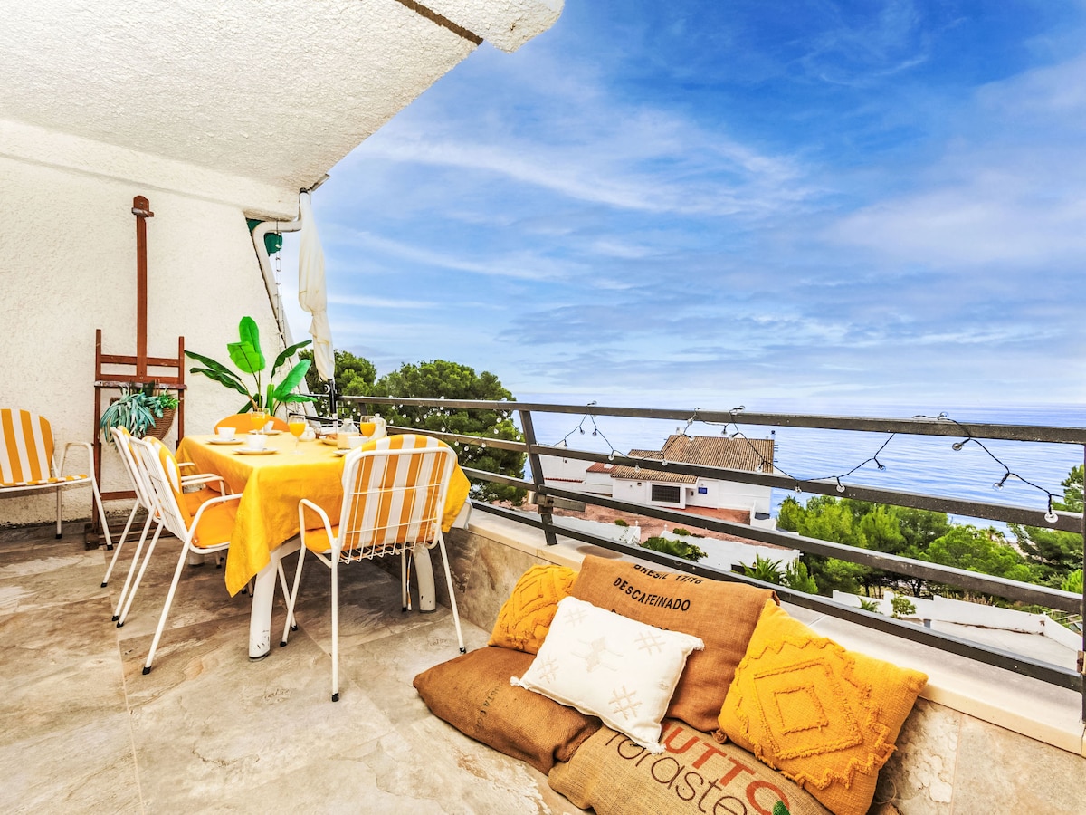 A balcony area is shown with a view of the sea and clear blue sky. A round dining table is set with yellow tableware, surrounded by four white chairs. Colorful cushions are placed on the floor, enhancing the outdoor space's comfort.