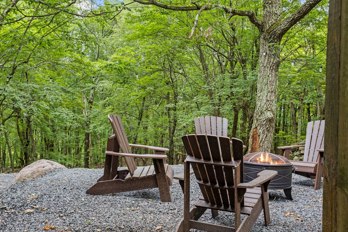Four wooden adirondack chairs are arranged around a fire pit in a gravel clearing, surrounded by lush green trees. The setting provides a natural atmosphere, ideal for outdoor gatherings and relaxing evenings by the fire.